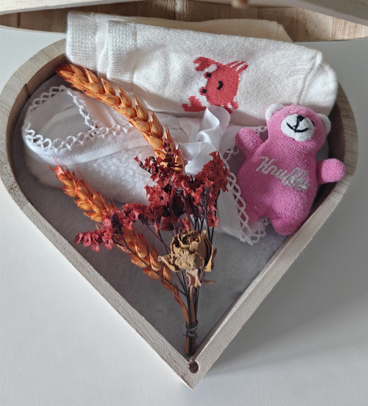 Een houten herinneringsdoosje in de vorm van een hart, met babysokjes, een doekje, een roze knuffeltje en droogbloemen
